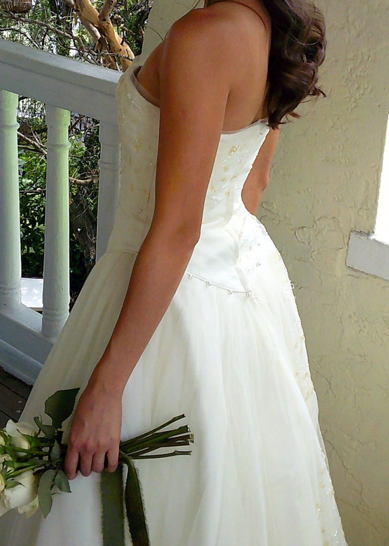 Peut inclure: Une robe bustier ivoire avec un corsage ajust&eacute; orn&eacute; de perles et de sequins d&eacute;licats, se prolongeant par une jupe en tulle superpos&eacute;e. Un fin cordon de petites perles blanches souligne la taille, accompagn&eacute; d'un bouquet de roses blanches.