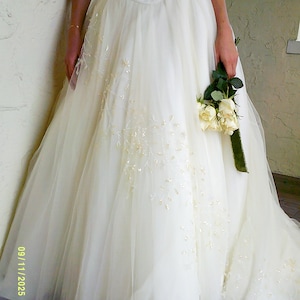 Peut inclure: Une robe longue couleur ivoire dot&eacute;e d'un corsage ajust&eacute; avec des perles complexes et une jupe en tulle volumineuse orn&eacute;e de perles et de sequins dispers&eacute;s. Le v&ecirc;tement est accompagn&eacute; d'un petit bouquet de roses couleur cr&egrave;me.