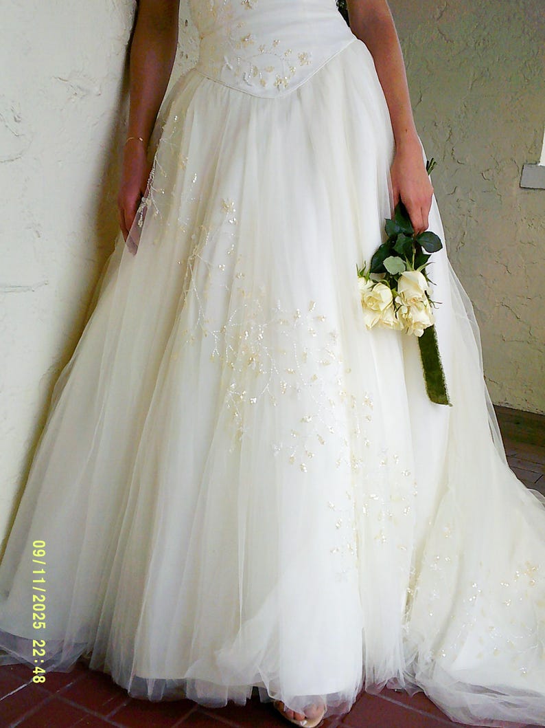 Peut inclure: Une robe longue couleur ivoire dot&eacute;e d'un corsage ajust&eacute; avec des perles complexes et une jupe en tulle volumineuse orn&eacute;e de perles et de sequins dispers&eacute;s. Le v&ecirc;tement est accompagn&eacute; d'un petit bouquet de roses couleur cr&egrave;me.