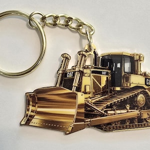 May include: A gold-toned metal keychain featuring a detailed illustration of a CAT D8T bulldozer. The heavy machinery design includes a large front blade, tracks, and the CAT logo printed on the side and front. A circular gold split ring is attached.