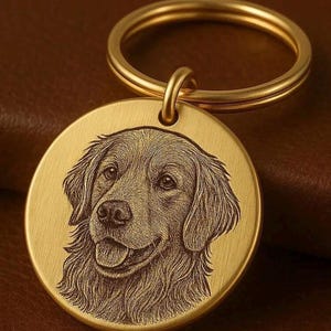 May include: A circular gold-toned metal keyring featuring a detailed laser-engraved portrait of a golden retriever dog. The pendant is attached to a matching gold-coloured split ring, showcasing a brushed metallic finish on the surface.