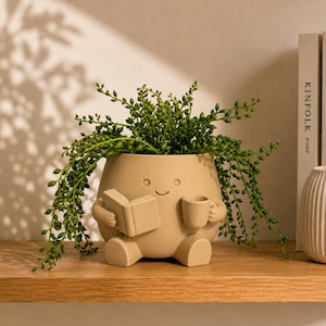 Könnte beinhalten: Ein matter beigefarbener Keramikübertopf in Form einer sitzenden Figur, die ein offenes Buch in der einen und eine kleine Tasse in der anderen Hand hält. Der Übertopf hat ein eingraviertes lächelndes Gesicht und eine grüne Erbsenpflanze.
