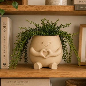 May include: A beige ceramic planter shaped like a rounded figure with a smiling face and arms forming a heart shape in front of its body. The planter contains a trailing green plant with small leaves that drape over the sides of the container.