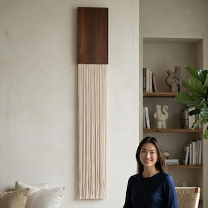 May include: A wall hanging featuring a rectangular dark brown wooden top section with a long, vertical fringe made of cream-coloured cotton cord hanging down from the base, creating a textured, linear decorative element on the wall.