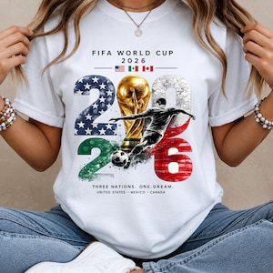 May include: A white t-shirt featuring a FIFA World Cup 2026 graphic. The design includes the text, the flags of the United States, Mexico, and Canada, a gold trophy, a soccer player, a soccer ball, and the large numbers 2026 with flag patterns.