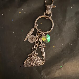 May include: A silver-toned metal keychain featuring a lobster clasp and a split ring. Attached are four charms: a textured feather, a small circular disc with a black line drawing, a faceted iridescent green heart, and a detailed moth pendant.