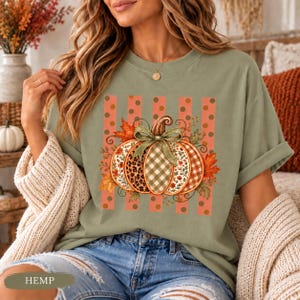 Op de afbeelding: Een saliegroen T-shirt met korte mouwen en een centrale pompoenafbeelding met segmenten in luipaardprint, groen gingham en bloemmotieven. De pompoen is gestrikt met een groen lint en omringd door herfstbladeren op koraalkleurige strepen.