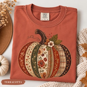 May include: A terracotta-colored t-shirt featuring a large graphic of a pumpkin composed of various patterned segments, including floral prints, plaid, and solid colors. The shirt has a Comfort Colors brand label at the neck with size L.