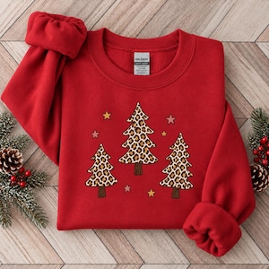 Könnte beinhalten: Ein leuchtend rotes Sweatshirt mit Rundhalsausschnitt und einer Grafik von drei Weihnachtsbäumen mit braun-schwarzem Leopardenmuster. Das Design enthält kleine, verstreute Sterne in Rosa, Gelb und Gold. Das Kleidungsstück hat ein Gildan-Etikett.