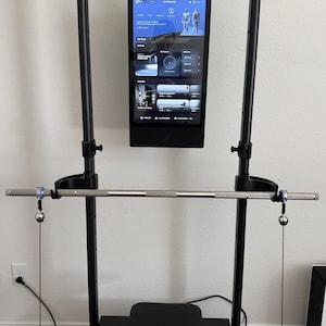 Può includere: Un attrezzo fitness domestico in metallo nero con telaio verticale e display touchscreen integrato. Lo schermo mostra un'interfaccia digitale con opzioni di allenamento. Un bilanciere argentato è fissato a cavi regolabili sul telaio.