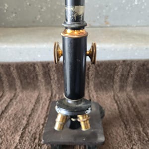 May include: A vintage black metal microscope featuring a cylindrical body, two brass adjustment knobs on the sides, and a rotating nosepiece with two brass objective lenses. The instrument is mounted on a square black base with a flat stage.