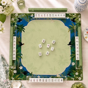 May include: A square mahjong game mat featuring a central light green area with a botanical print, surrounded by a blue border with swallows and palm leaves. Four clear acrylic tile racks hold white mahjong tiles with red and black characters.
