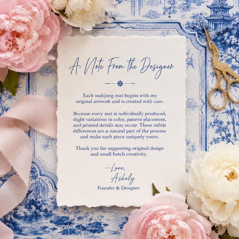 May include: A rectangular card with deckled edges featuring blue printed text titled A Note From The Designer. The card explains the production of mahjong mats and is signed by Ashely. It is surrounded by pink peonies, a silk ribbon, and gold scissors.
