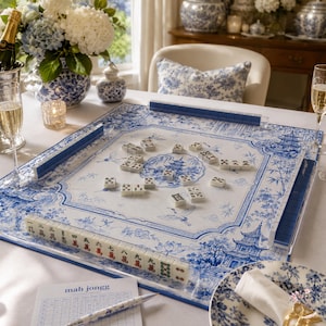 May include: A square mahjong game board featuring a blue and white chinoiserie pattern with a central pagoda illustration. The board includes clear acrylic tile racks and white mahjong tiles with black and red characters, set on a white tablecloth.