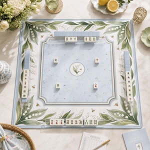 May include: A square mahjong mat featuring a light blue center with a lily of the valley illustration and four tiles, surrounded by a border of green lily of the valley leaves and small bees. Four tile racks hold rows of mahjong tiles.