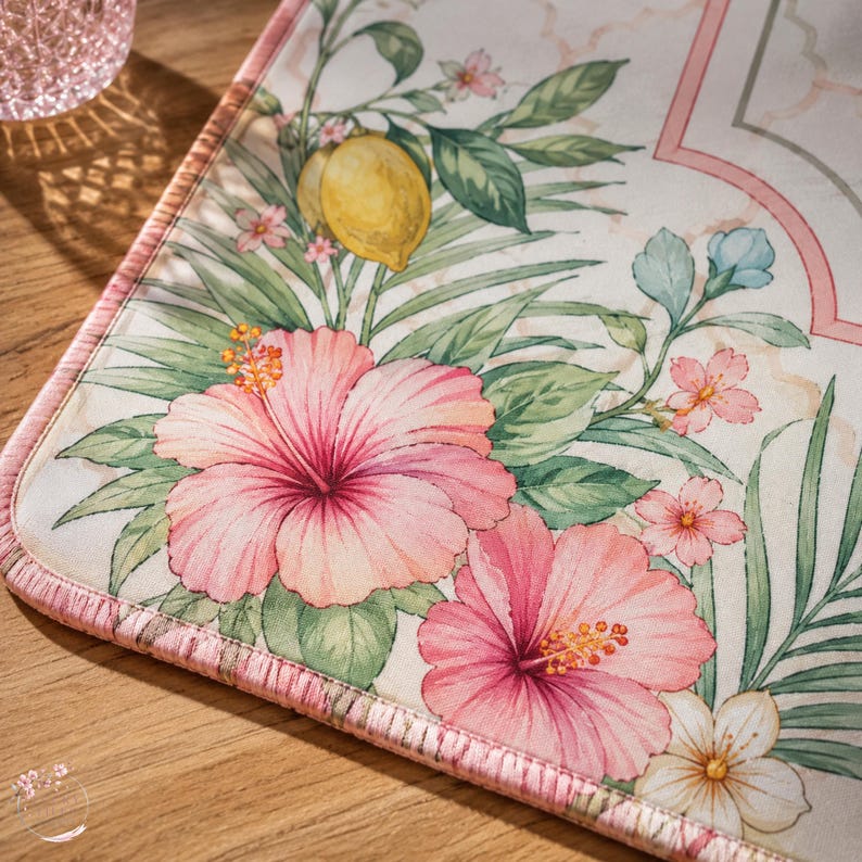 May include: A rectangular fabric mat featuring a botanical print with large pink hibiscus flowers, green palm leaves, a yellow lemon, and smaller light pink and white blossoms. The mat has a finished pink stitched edge and a light cream background.