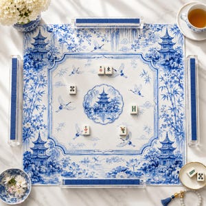 May include: A square mahjong mat featuring a blue and white chinoiserie pattern with pagodas, cranes, and floral motifs. The mat includes four clear acrylic tile racks containing blue tiles and five scattered mahjong tiles on the surface.