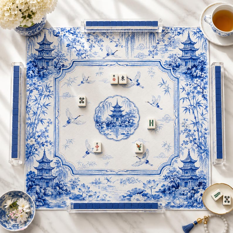May include: A square mahjong mat featuring a blue and white chinoiserie pattern with pagodas, cranes, and floral motifs. The mat includes four clear acrylic tile racks containing blue tiles and five scattered mahjong tiles on the surface.