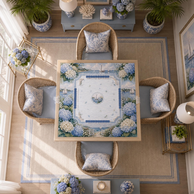 Puede incluir: Una mesa de juego cuadrada con un tablero de tem&aacute;tica costera, bordes de flores de hortensia azules, veleros y un motivo n&aacute;utico central, rodeada por cuatro sillas de rat&aacute;n trenzado con cojines azul claro y almohadones.