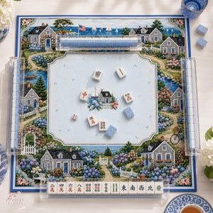 Puede incluir: Un juego de Mahjong que incluye un tablero cuadrado con una ilustraci&oacute;n de un paisaje de casas costeras, fichas azules y blancas, y soportes de acr&iacute;lico transparente. El tablero muestra casas, jardines y un velero. Las fichas tienen caracteres chinos.