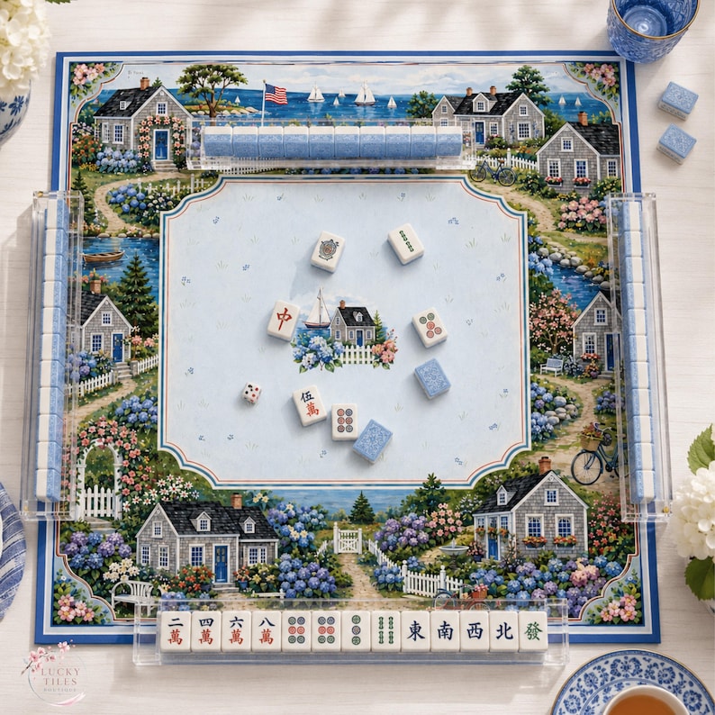 Puede incluir: Un juego de Mahjong que incluye un tablero cuadrado con una ilustraci&oacute;n de un paisaje de casas costeras, fichas azules y blancas, y soportes de acr&iacute;lico transparente. El tablero muestra casas, jardines y un velero. Las fichas tienen caracteres chinos.