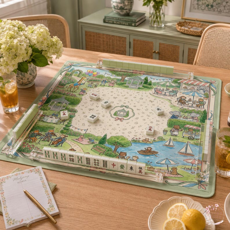 May include: A square mahjong mat featuring a colorful park illustration with animals, a pond, and a gazebo. The mat includes clear acrylic tile racks on all four sides, with white mahjong tiles placed on the surface and within the racks.