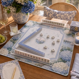Puede incluir: Un juego de Mahjong que incluye un tapete rectangular con estampado floral de hortensias azules y blancas, una ilustraci&oacute;n n&aacute;utica central y fichas blancas con caracteres negros y rojos. Incluye un soporte transparente y una tarjeta de puntuaci&oacute;n.