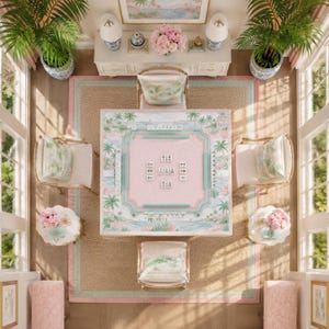 May include: A square mahjong table featuring a pastel pink and green tropical palm tree print, surrounded by four upholstered chairs with matching cushions, set on a large woven area rug in a room with two potted palm plants and side tables.