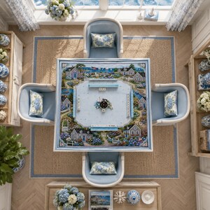 Puede incluir: Una mesa de mahjong cuadrada con una superficie azul claro que muestra una ilustraci&oacute;n de un pueblo costero, rodeada por cuatro sillas tapizadas en azul claro con cojines florales, sobre una alfombra tejida en una habitaci&oacute;n decorada.