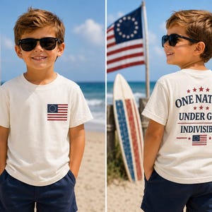 May include: A cream-colored short-sleeve t-shirt featuring a small American flag graphic on the left chest and a large back print that reads ONE NATION UNDER GOD INDIVISIBLE with a small American flag graphic centered below the text.