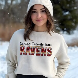 May include: A cream-colored crewneck sweatshirt featuring the black text Santa's Favorite Team above the word RAVENS in a red and black buffalo plaid pattern with a leopard print border, paired with a white knit beanie.
