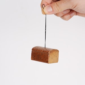 May include: A small, brown loaf of bread with a textured surface, hanging from a black string. The bread has a rectangular shape with a rounded top and side details. The bread is suspended against a white backdrop.