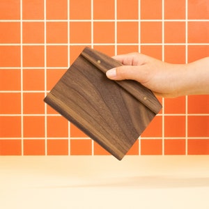 May include: A dark brown wooden clipboard with brass accents. The clipboard is held in a hand against a background of orange tiles.