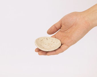 May include: A small, speckled ceramic dish in a light beige color. The dish is oval-shaped and held in a person's hand against a white background. The dish has small, dark brown speckles throughout.