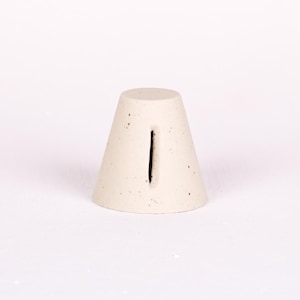 May include: A cream-colored, cone-shaped object with a vertical black slit. The object has a speckled texture and a flat top. The design is minimalist and modern, suitable for home decor or as a unique accent piece.