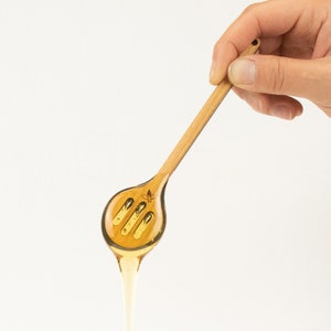 May include: A wooden honey dipper with a bee design. The dipper is filled with honey and dripping down the handle.