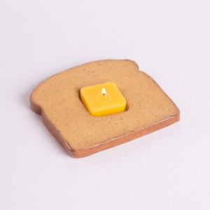 May include: A ceramic candle holder shaped like a slice of toast. The toast-shaped holder is tan with brown edges and speckled details. A square yellow candle sits in a recessed area, with a lit flame.