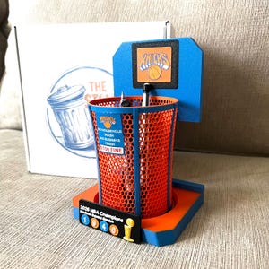 May include: A blue and orange mesh pencil holder shaped like a basketball hoop with a backboard featuring the Knicks logo. The holder includes a sign reading NO HOUSEHOLD TRASH NO BUSINESS TRASH $100 FINE and a 2026 NBA Champions Madison Square Garden plaque.