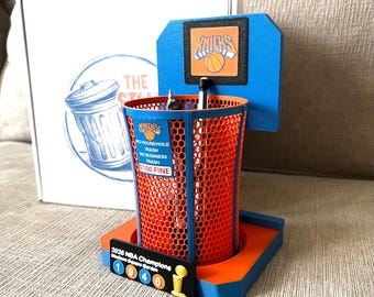 Peut inclure: Un porte-crayon en maille bleue et orange en forme de panier de basket avec un panneau affichant le logo Knicks. Il porte l'inscription NO HOUSEHOLD TRASH NO BUSINESS TRASH $100 FINE et une plaque 2026 NBA Champions Madison Square Garden.
