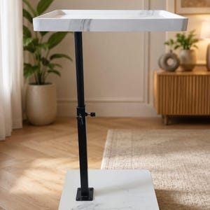 May include: A mobile side table featuring a white faux marble rectangular top and a matching white faux marble base. The two surfaces are connected by a single vertical black metal pole with an adjustable height mechanism and a black knob.