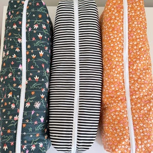 May include: Three rectangular quilted fabric pouches with white zippers. The left pouch is dark green with a floral pattern, the center pouch features black and white horizontal stripes, and the right pouch is orange with a small white floral print.