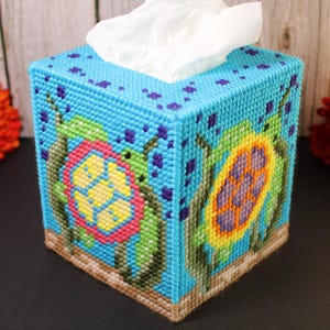 May include: A square tissue box cover crafted from blue plastic canvas with a cross-stitch design featuring two sea turtles with colourful shells, green seaweed, and dark blue bubbles. The base is tan, and white tissues protrude from the top.
