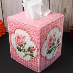 May include: A square tissue box cover crafted from pink plastic canvas with a grid texture. Each visible side features a white, cloud-shaped inset displaying a cross-stitched design of a pink rose with green leaves and a brown stem.