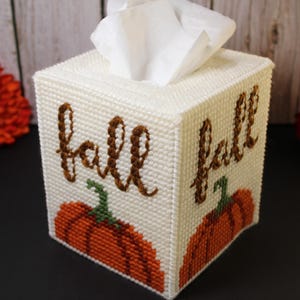 Peut inclure: Une boîte à mouchoirs carrée en toile plastique blanche avec le mot fall brodé en fil marron au-dessus d'un motif de citrouille orange avec une tige verte sur deux côtés visibles. La housse est faite d'un matériau en forme de grille.