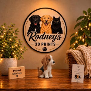 May include: A circular wall sign displaying illustrations of a black dog, a yellow dog, and a black cat with the text Rodney's 3D PRINTS and a paw print. Below is a small crocheted beagle-style dog figurine and a wooden block with the text Printed with love.