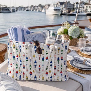 May include: A white tote bag featuring a repeating pattern of colorful nautical buoys and small anchors. The bag has thick white rope handles and contains a blue and white striped towel, a book titled Coastal Living, sunglasses, and sunscreen.