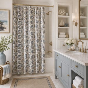 May include: A shower curtain featuring a repeating pattern of blue and tan oyster shells and seaweed. The curtain hangs from a brass rod in a bathroom with a gray vanity, a marble countertop, a woven rug, and built-in shelving with decor.