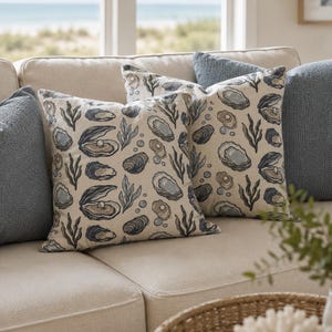 May include: Two square throw pillows featuring a cream-colored fabric with a repeating pattern of dark blue and grey oysters, some containing pearls, interspersed with seaweed and bubbles, flanked by two solid textured slate blue throw pillows.