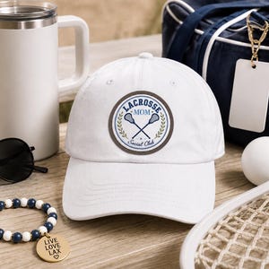 Peut inclure: Une casquette de baseball blanche ornée d'un écusson circulaire portant l'inscription "LACROSSE MOM" et "Social Club" en bleu, accompagnée d'un motif de deux crosses de crosse croisées et de branches de laurier vertes sur le devant.