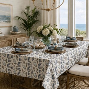 Puede incluir: Una mesa de comedor rectangular cubierta con un mantel blanco que presenta un patrón repetitivo azul y gris de conchas de ostra y algas. La mesa está dispuesta con manteles individuales trenzados, platos blancos y cristalería.